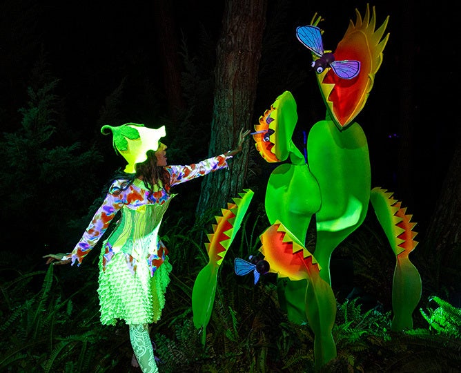 Actor dressed in a neon fairy outfit interacting with a  large scale Venus flytrap sculpture