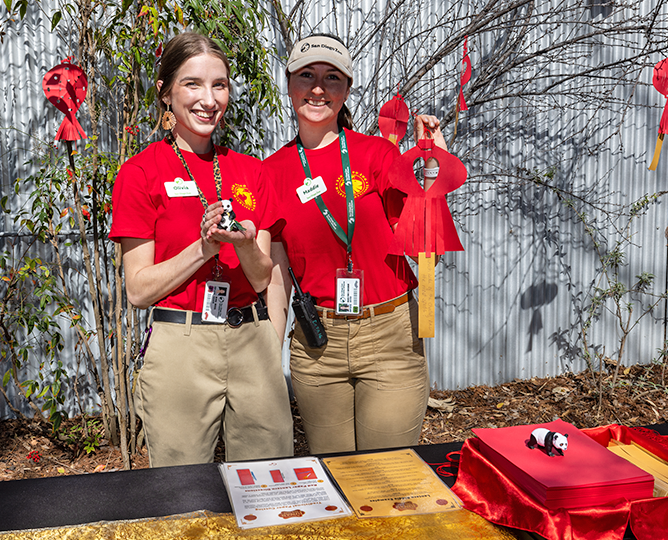 A pair of young people in bright red shirts holding paper lanterns behind craft table