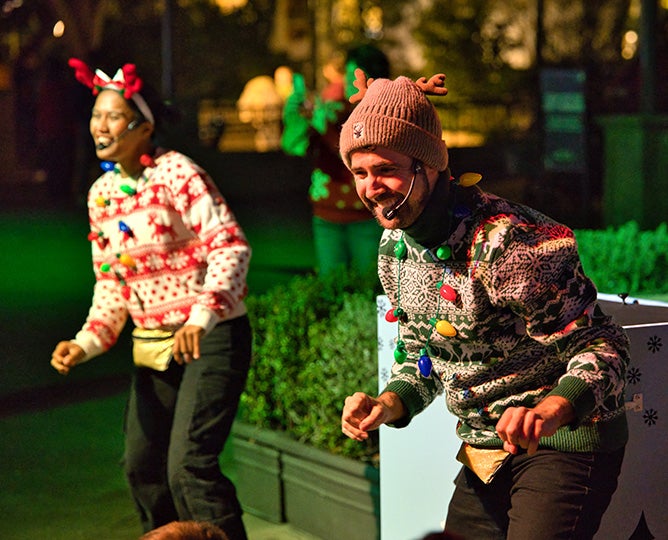 performers dressed in holiday sweaters with headset talk excitedly to a crowd